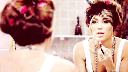gloss-miley-cyrus Miley Cyrus com redinha no cabelo, passando gloss labial frente a espelho com regata branca.