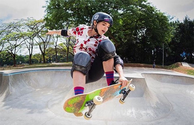 A imagem mostra uma menina branca, vestindo equipamento de proteção preto (capacete, joelheira e cutuveleira), fazendo uma manobra de skate numa pista de skate.