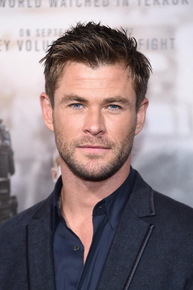 Chris Hemsworth posando para foto; ele está com terno preto e camisa azul-escuro com expressão neutra