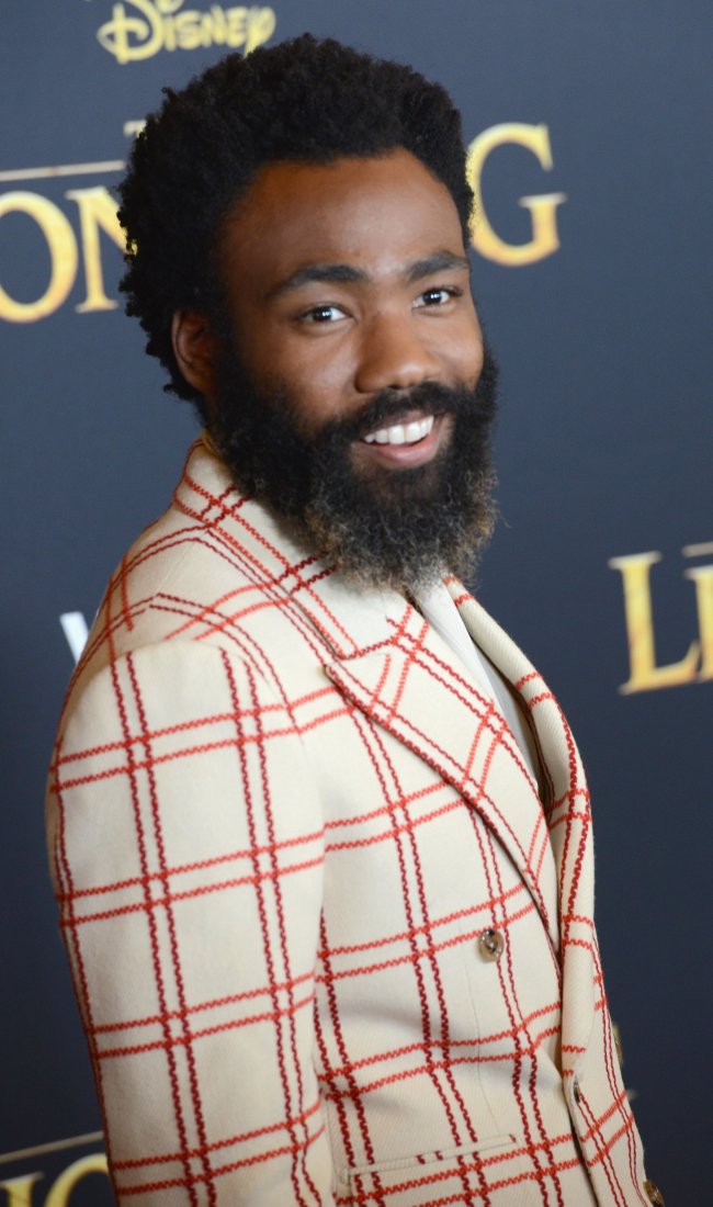 donald-glover-o-rei-leao-premiere Donald Glover na premiere de O Rei Leão; ele está posando sorrindo de lado no carpet e usa um paletó branco com detalhes vermelhos quadriculados