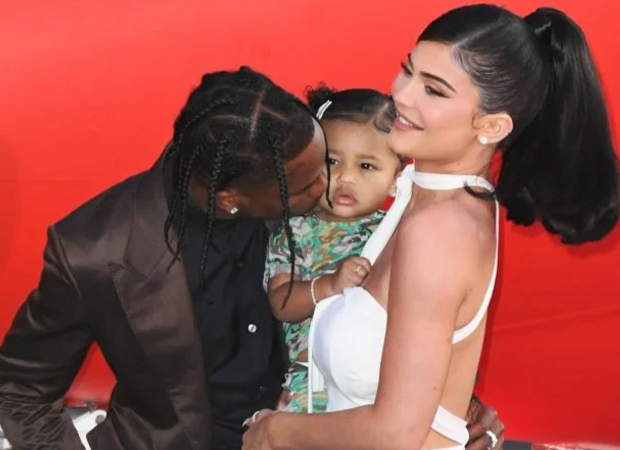 Travis Scott com Kylie Jenner e Stomie; os três estão posando no carpet de premiação e Travis está dando um beijo na bochecha de Stormi, que está no colo de Kylie; a empresária sorri para as câmeras enquanto segura a filha