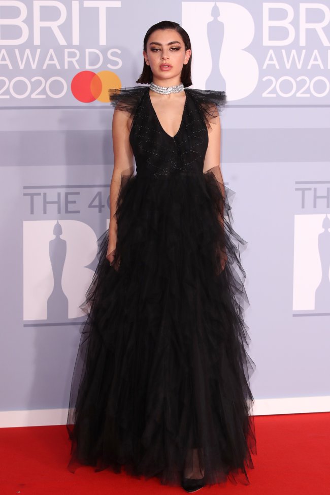 Charli XCX posando para foto no red carpet do BRITs 2020 usando um vestido preto com saia volumosa e um colar brilhante prata