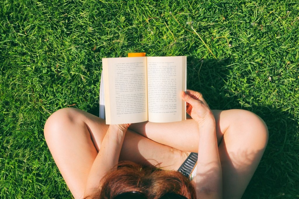 Imagem de uma mulher branca lendo um livro em um gramado.