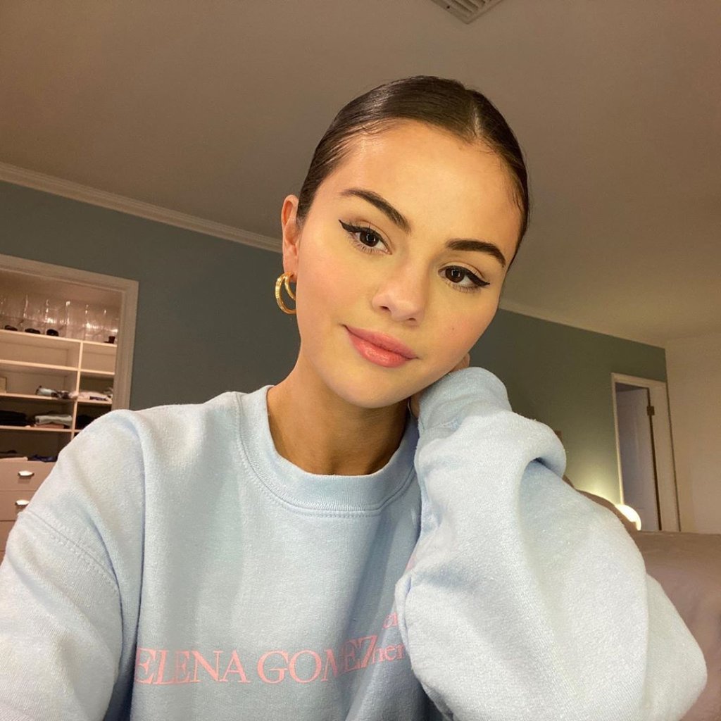 Selfie de Selena Gomez com o cabelo preso, usando moletom branco e sorrindo levemente para câmera com o rosto apoiando em uma das mãos