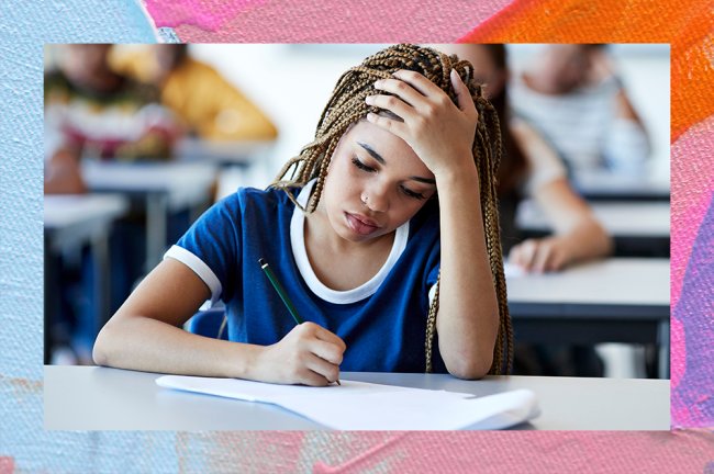 A imagem mostra uma menina negra, com o cabelo longo e escuro em tranças. Ela está usando o que parece ser um uniforme escolar, com uma camiseta azul com a ponta das mangas e a gola brancas. Ela está sentada em uma mesa, escrevendo em uma folha com uma mão enquanto a outra apoia a cabeça, como se estivesse pensando. No fundo, mesmo que levemente desfocado, é possível ver outras carteiras e pessoas atrás dela, como se estivesse numa escola ou numa sala de prova.