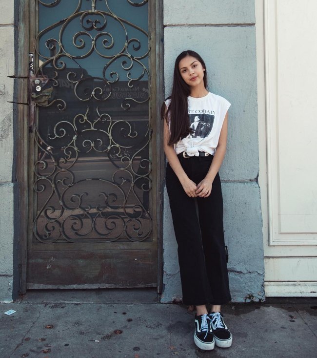 Olivia Rodrigo usando calça e tênis pretos, camiseta branca com estampa preta e nó na região da barriga