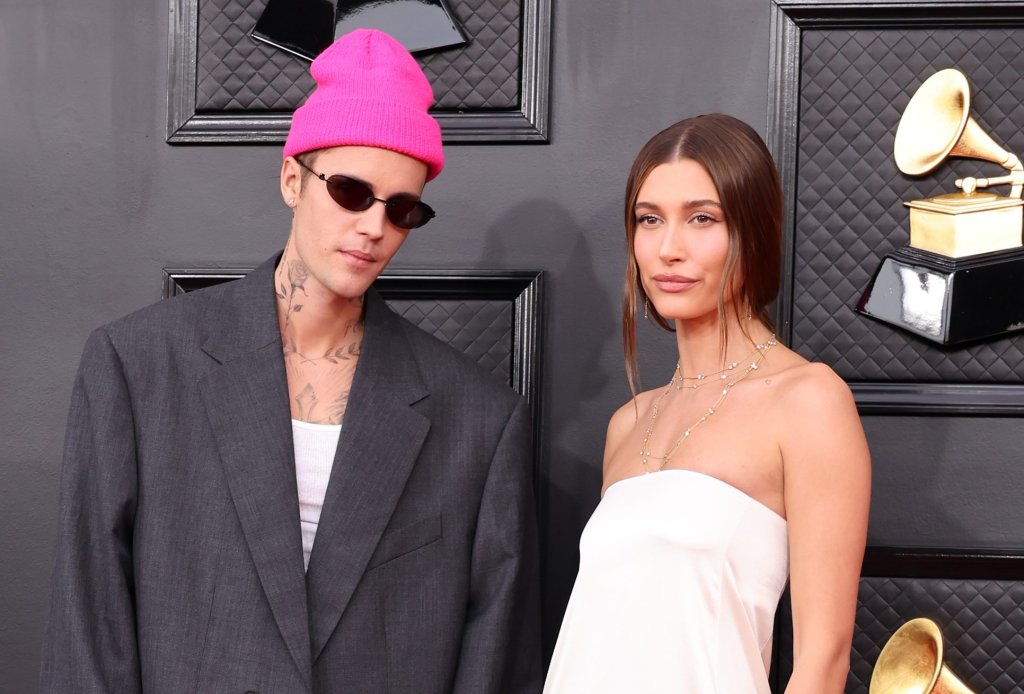 Imagem mostra Justin Bieber e Hailey Bieber posando juntos no tapete vermelho da 64ª edição do Grammy Awards. Justin usa óculos escuro, gorro rosa pink e um blazer e calça cinza escura com modelagem ampla e oversized. Enquanto Hailey um vestido branco de cetim sem alças com modelagem mais reta. Ambos olham para a câmera com expressão leve.