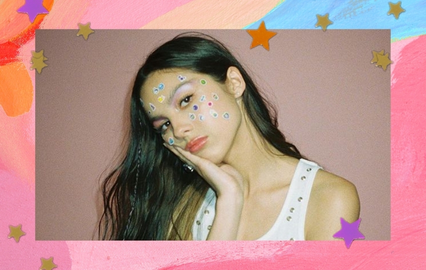 Olivia Rodrigo com adesivos colados no rosto, que está apoiando em sua mão; a margem é uma textura em tons de rosa, laranja e azul com estrelas amarelas, laranjas e lilás como decoração