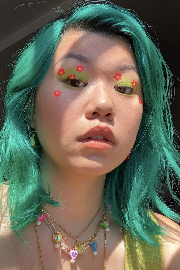 Maquiagem estilo kidcore Inspiração de maquiagem colorida com flores de cor laranja e sombra esfumada verde, jovem com cabelo verde água posando para foto com close em seu rosto.