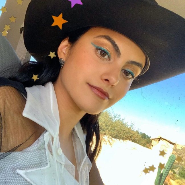 Camila Mendes Atriz Camila Mendes com delineado verde, usando chapéu de cowboy preto, camisa branca com babado e posando para selfie com uma expressão tranquila