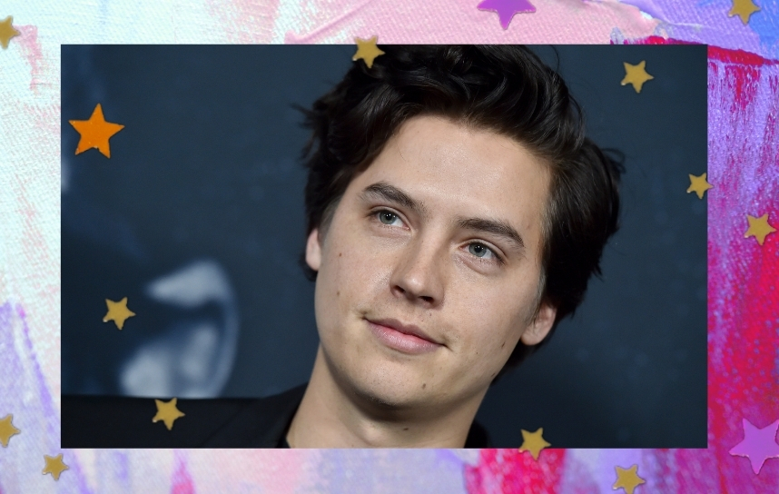 Ator Cole Sprouse, na imagem só conseguimos ver do seu pescoço para cima, ator usa blazer preto e tem expressão sorridente.