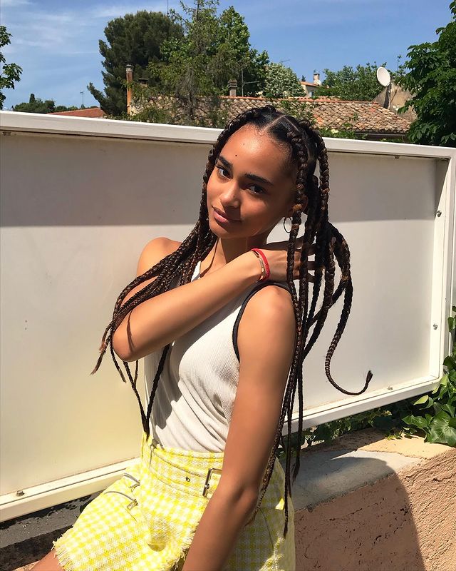 Mélanie do Now United com trancinhas Mélanie, do Now United, com trancinhas no cabelo. Usando regata branca e saia amarela xadrez com a barra desfiada.