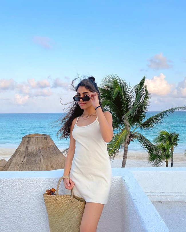 Nour, do Now United, com o cabelo meio preso Nour, do Now United, com o cabelo meio preso, com óculos escuros, posando para foto apoiada em um muro baixo, com vestido de alças finas branco, segurando uma bolsa de palha, ao fundo se vê: o céu azul, o mar azul, alguns coqueiros e a areia.