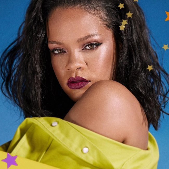 Rihanna Cantora Rihanna posando para foto com delineado gatinho, batom vermelho fechado, com expressão séria, de lado, com a camisa verde caída em seu ombro.
