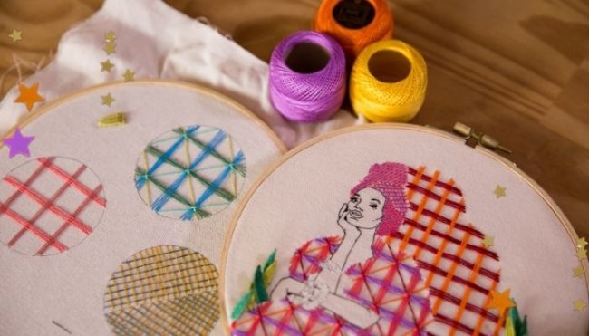 bordados Mesa de madeira com bastidores brancos e linhas laranja, amarela e roxa posicionadas. Nos bastidores vemos bordados coloridos nas cores rosa, laranja, verde e azul, em um deles tem o desenho de uma mulher sentada com a mão apoiada no rosto com uma expressão pensativa e usando turbante na cabeça.