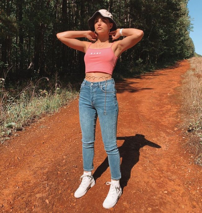 Garota usando top cropped rosa, bucket hat, calça jeans skinny e coturno branco, além de cinto de correntes em formato de coração na barriga. Ela está em pé com os dois braços dobrados perto da nuca.