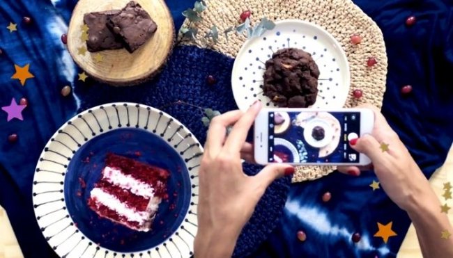 fotografia instagram Mesa com toalha azul, com pratos, e bolos, sendo eles: red velvet, cupcake de chocolate e brownie. Também conseguimos ver duas mãos segurando um celular posicionado para fotografar a composição da mesa.