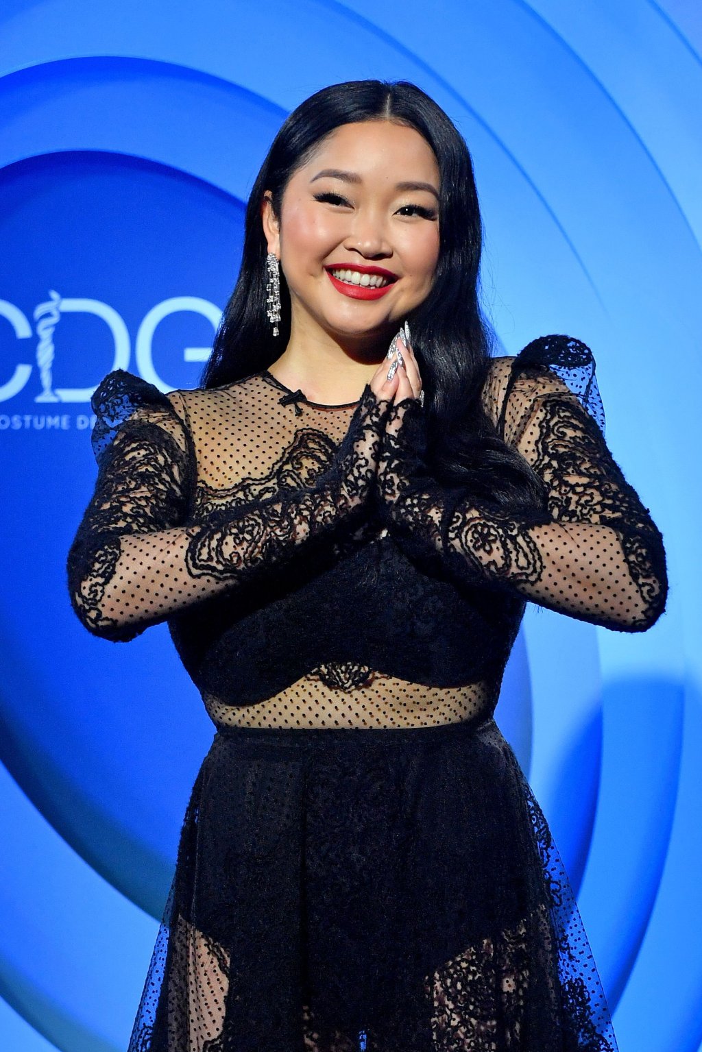 Lana Condor está usando um vestido preto de renda com transparências. A foto está aproximada e pega apenas da cintura para cima. Ela está usando batom vermelho, sorrindo com o olhar levemente para o lado e as duas palmas das mãos estão juntas na altura do peito.