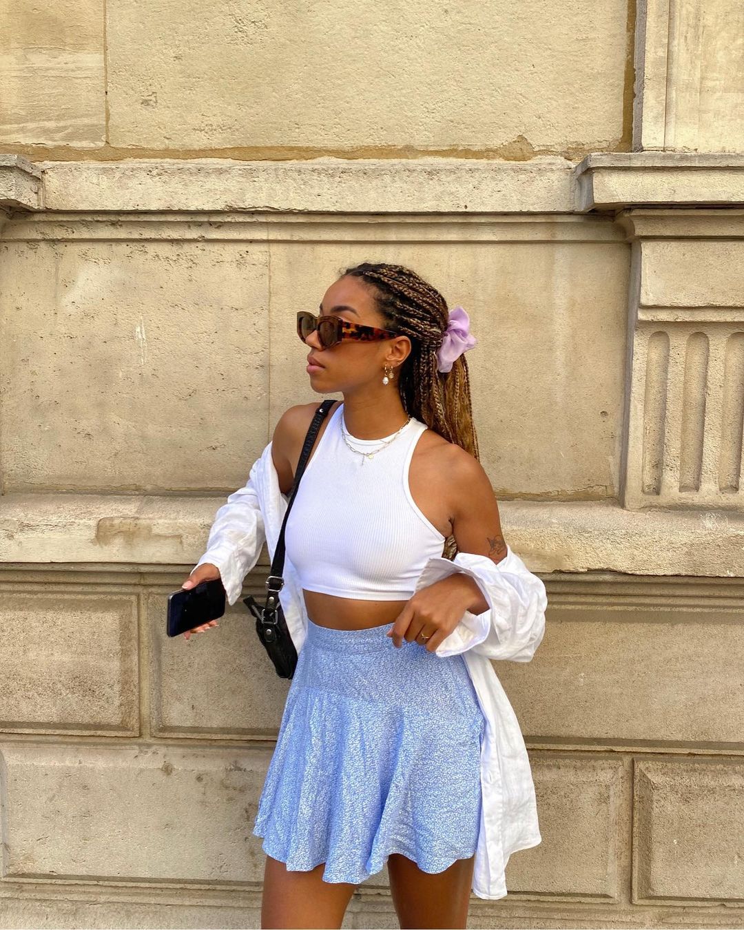 Garota usando top cropped branco com saia rodada azul e camisa branca por cima. Ela está com uma bolsa preta pendurada em um dos ombros, segurando o celular em uma das mãos, cabelo preso em um rabo com scrunchie lilás e óculos de sol. Ela está olhando para o lado.