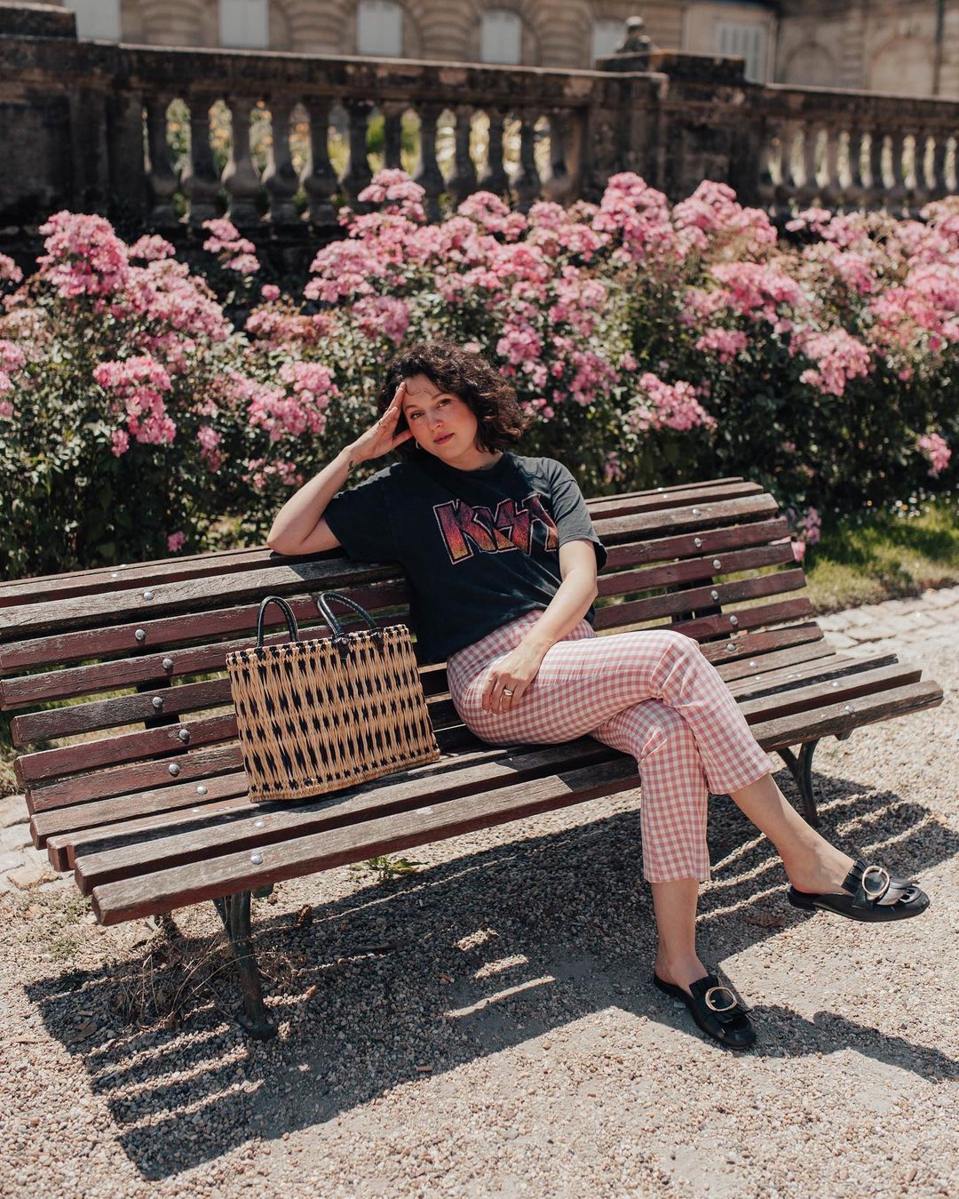 look-xadrez-vichy-anaiswho Garota usando look com calça xadrez vichy vermelha, camiseta preta da banda Kiss e oxford preto. Ela está sentada em um banco marrom, com uma das mãos apoiadas na coxa, e a outra na cabeça. Ao lado dela, tem uma maxibolsa de palha com detalhes em preto. Sua expressão facial é séria.