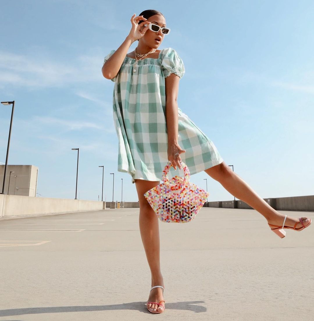 look-xadrez-vichy-byaimeekelly Garota usando look com vestido xadrez vichy verde e sandália branca e vermelha de tiras com saltinho. Com uma das mãos, ela segura os óculos de sol brancos, e com a outra uma bolsa de miçangas coloridas. Uma das pernas está levantada para o lado e sua expressão facial é de surpresa, com a boca aberta.