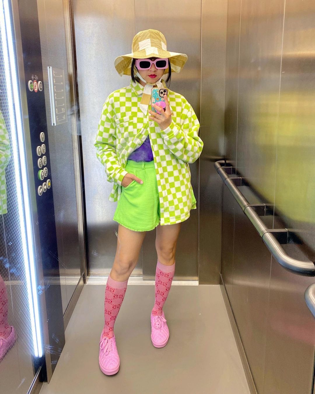 Garota usando casaco xadrez vichy verde com short verde neon, meia rosa até o joelho e tênis rosa, além de um chapéu xadrez vichy amarelo. Ela está em pé, em frente a um espelho, usando óculos de sol cor-de-rosa, com uma das mãos no bolso, e a outra segurando o celular para tirar a foto em frente ao espelho.