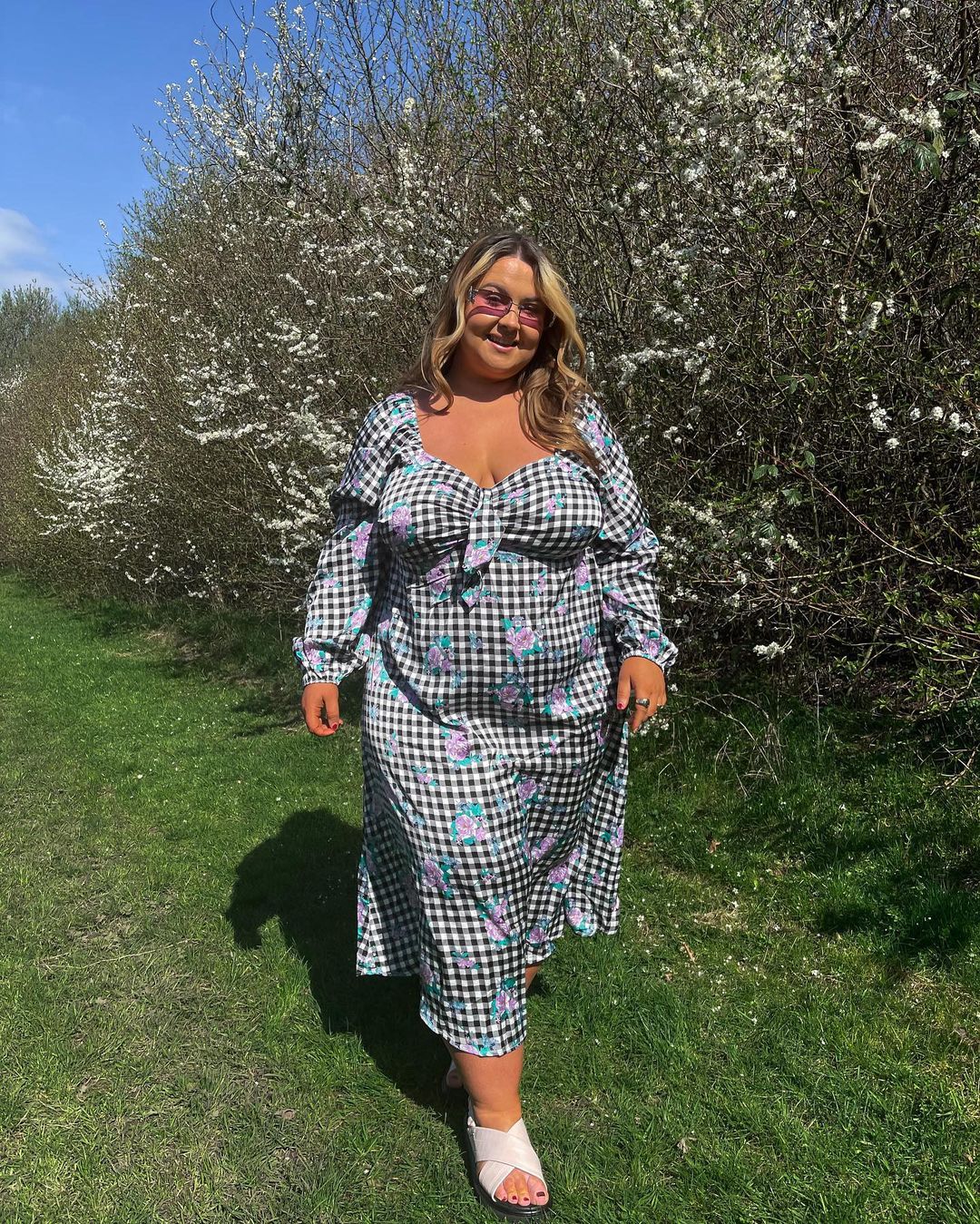 look-xadrez-vichy-is0belx Garota usando look com vestido longo xadrez vichy preto e branco e algumas flores em rosa. Ela usa uma sandália branca, óculos de sol com lentes rosas, está em pé e sorrindo.