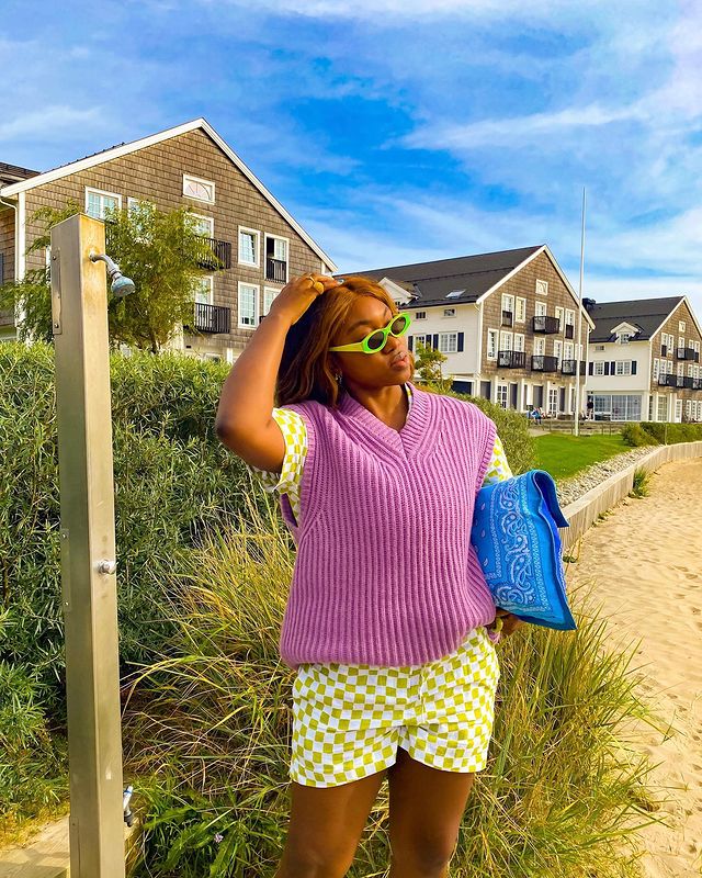 Garota usando conjuntinho de camisa de manga curta e short verdes com estampa xadrez vichy. Por cima da camisa, um colete de tricô lilás. Ela usa óculos de sol em verde neon, carrega uma bolsa azul com estampa de bandana embaixo de um dos braços, e a outra mão está mexendo na parte de cima do cabelo. Sua cabeça está levemente inclinada para cima e sua expressão facial é séria.