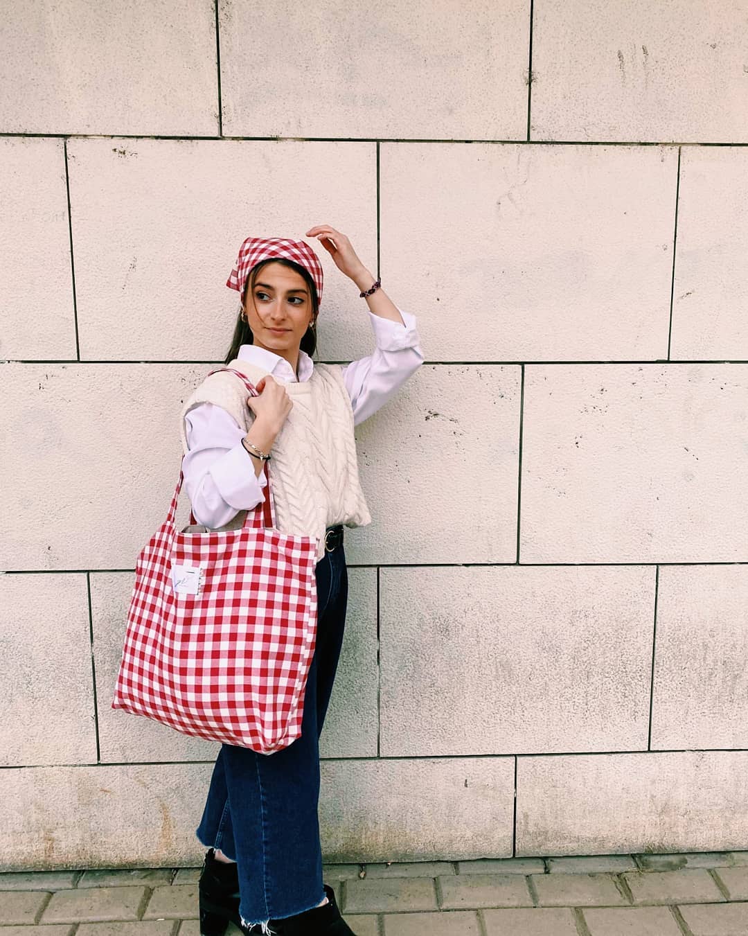 look-xadrez-vichy-sewithyai Garota usando look com camisa branca e suéter de tricô off-white por cima, calça jeans de lavagem escura e bota preta. Ela está com uma maxibolsa pendurada em um dos ombros que tem estampa xadrez vichy vermelha, assim como o lenço na sua cabeça. Uma das mãos segura a alça da bolsa, enquanto a outra está perto do lenço. Ela está com a cabeça inclinada para o lado.