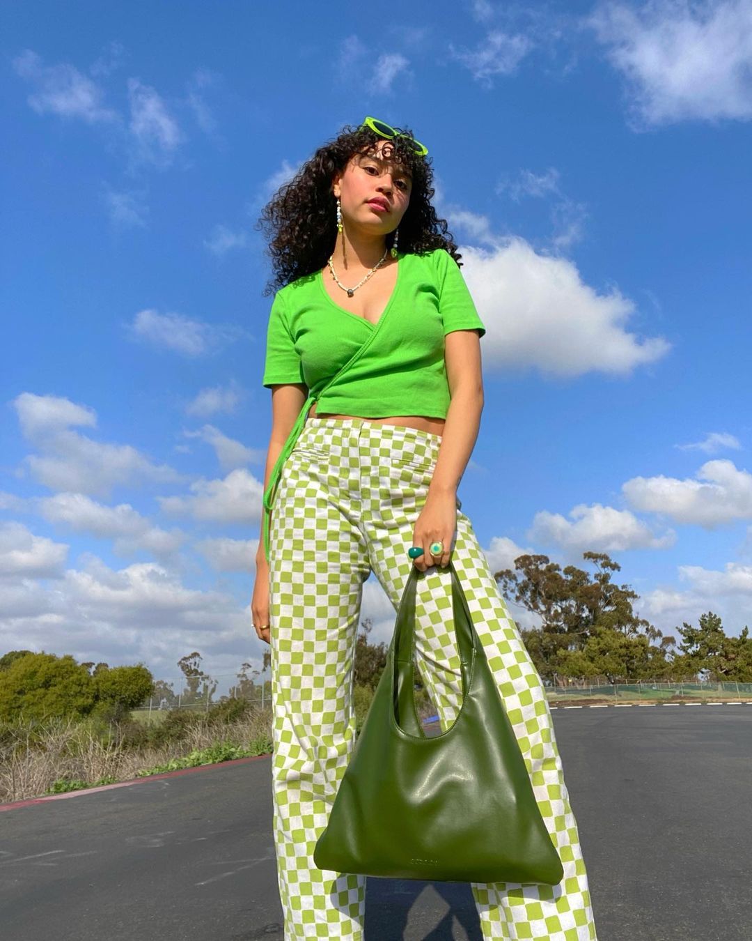 look-xadrez-vichy-thatcurlytop- Garota usando blusa verde neon e calça xadrez vichy verde de cintura alta. Ela está em pé, com os óculos de sol verdes neon na cabeça, uma das mãos segurando uma maxibolsa verde escura. Ela está em pé, olhando para baixo, em direção à câmera.