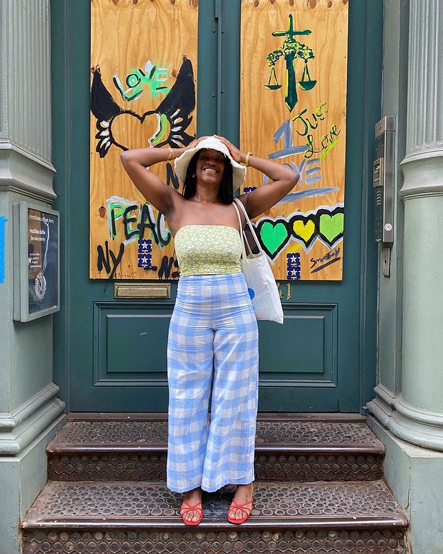 look-xadrez-vichy-thelanguageofyolande Garota usando top cropped sem alças verde com estampa floral e calça pantalona com estampa xadrez azul vichy. Ela ainda usa uma sandália vermelha, um chapéu bege e uma ecobag branca pendurada em um dos ombros. As duas mãos estão na cabeça e ela está sorrindo.