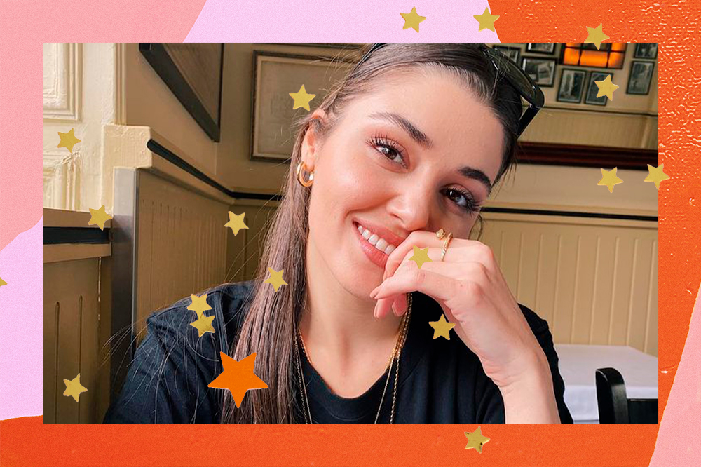 Montagem com foto da atriz Hande Erçel. Ela está sorrindo com uma das mãos perto da boca e usa uma camiseta preta e um óculos de sol preto no topo da cabeça. Na montagem, há um fundo rosa e laranja e várias estrelas douradas pela foto.