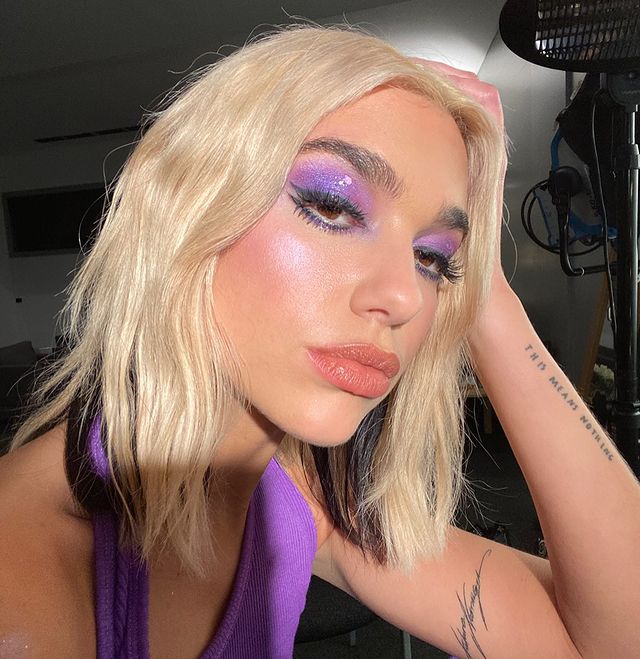 maquiagem-dua-lipa-08 Dua Lipa usando maquiagem com sobra roxa com efeito glossy. Ela está com uma regata no mesmo tom de roxo. Uma das mãos está apoiada na cabeça e sua expressão facial é séria.