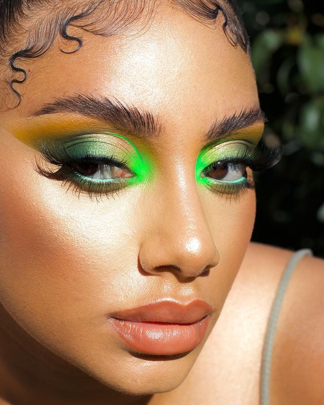 Garota usando maquiagem com diferentes tonalidades de verde nos olhos. A pele está bem iluminada, com bronzer, batom cor de boca e cílios postiços. Na foto, só dá para ver o rosto dela. Sua expressão é séria e ela olha para o lado.