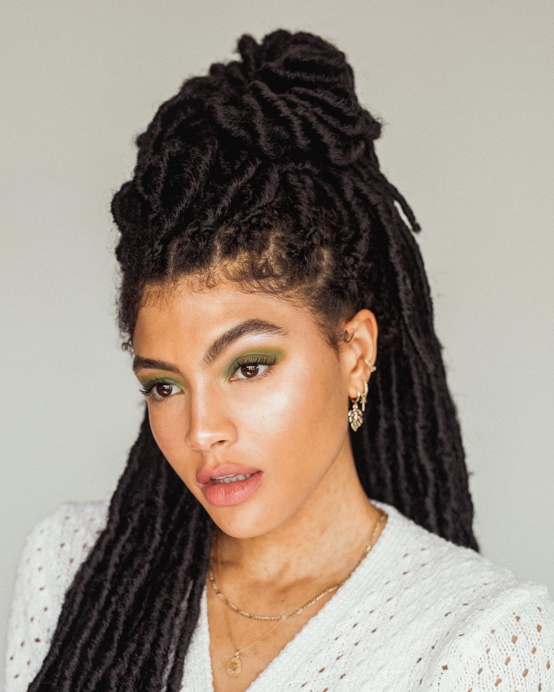 Garota usando maquiagem com sombra verde oliva e suéter branco. Ela está olhando para baixo, com expressão facial séria e a boca levemente aberta. Seu cabelo está com box braids, e a foto só mostra dos ombros para cima.