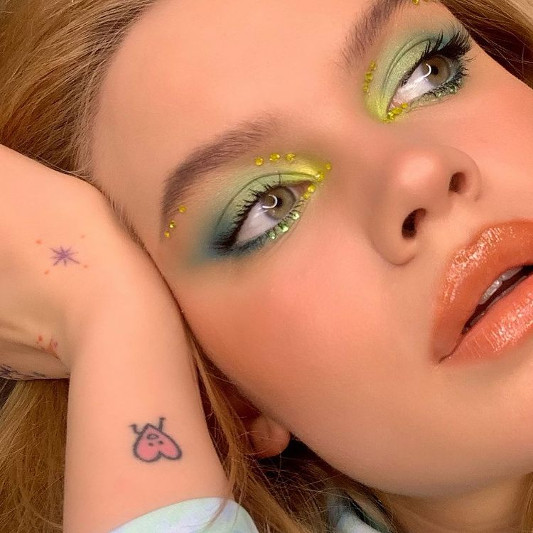 maquiagem-olho-verde-weepeace Garota usando maquiagem com diferentes tons de verde. Na foto, só dá para ver seu rosto e uma mão apoiada na cabeça. Ela olha para o lado e a boca está um pouco aberta.