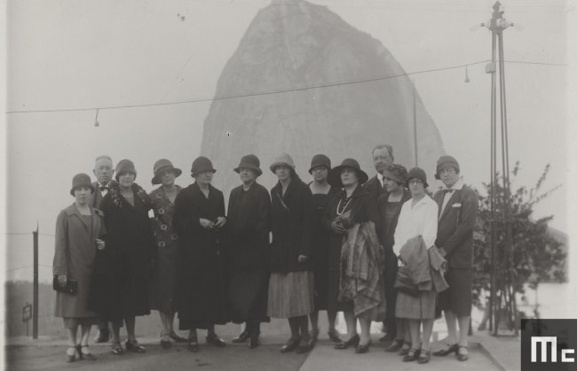 Foto em preto e branco da cientista Marie Curie durante visita ao Brasil; ela está rodeada de pessoas e o Pão de Açúcar aparece ao fundo