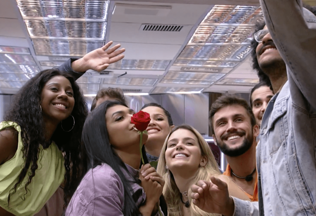 João segura um celular acima de sua cabeça. Camilla, Caio, Pocah, Juliette, Viih Tube, Fiuk e Arthur estão juntos com ele. olhando para a câmera e sorrindo. Pocah segura uma rosa vermelha encostada em sua boca