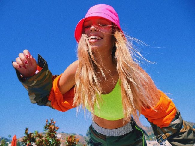 Salem Ilese do hit mad at disney Cantora Salem Ilese, do hit Mad at Disney, posando para foto com cropped verde florescente, blusa de frio laranja com estampa do exército e bucket hat rosa