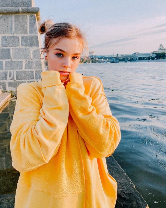 sofya-now-united Sofya, do grupo Now United, usando blusa de moletom amarela e com coquinhos laterais em sua cabeça posando para foto com as duas mãos apoiadas nas laterais inferiores de seu rosto.