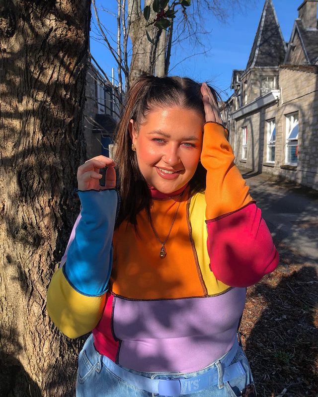 Garota usando blusa de patchwork azul, amarela, laranja, rosa e lilás. Ela está sorrindo, com uma das mãos no cabelo, e a outra levantada perto do rosto.