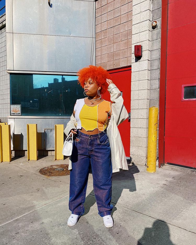 tendencia-patchwork-finessabuxtable Garota usando blusa cropped branca, laranja e amarela de patchwork com calça jeans baggy, tênis branco e casaco bege. Ela está em pé, com uma das mãos no cabelo crespo vermelho e, a outra, segurando uma bolsa branca pequena. Sua expressão facial é séria e ela está olhando para o lado.