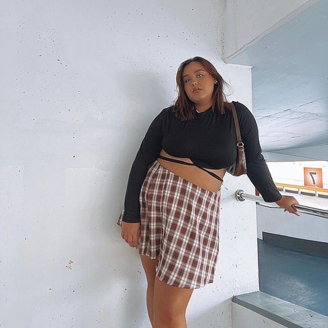 tendencias-outono-inverno-jadagiddings Garota está usando blusa de manga longa cropped preta com saia xadrez marrom e off-white e uma bolsa baguete pendurada em um dos ombros. Ela está com o corpo inclinado e uma das mãos apoiadas em um corrimão.