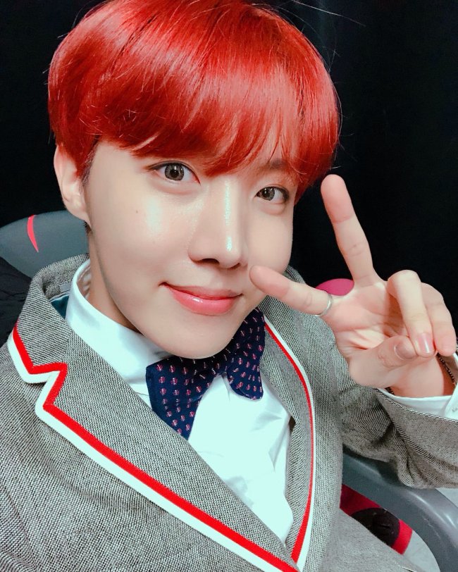 BTS J-Hope com cabelo colorido J-hope, do grupo BTS, com cabelo colorido, fazendo sinal de paz e amor, usando gravata borboleta e blazer cinza com lista vermelha e branca.