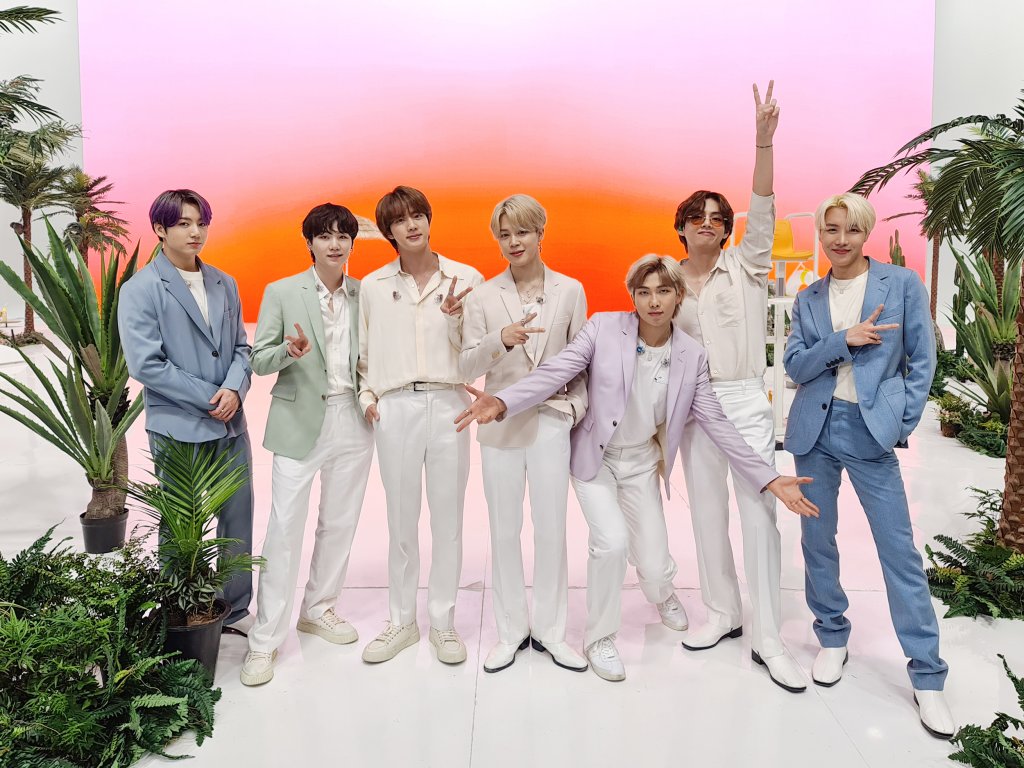 Integrantes do BTS posando para foto; eles estão alinhados na horizontal em um fundo colorido com tons de rosa e laranja; os dois integrantes de cada ponta da fileira usam roupas azuis enquanto o restante está vestindo trajes brancos