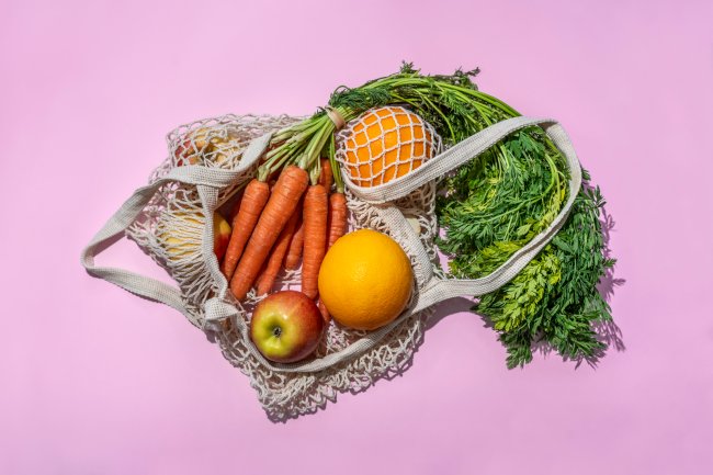 Sacola de feira ecológica cheia de frutas e legumes dentro. O fundo é rosa.