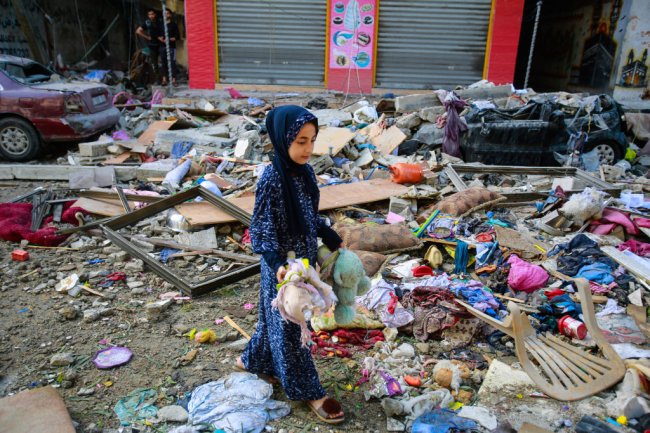 Uma garotinha está no meio de escombros, em uma rua de Gaza, com o que restou de alguns ursinhos de pelúcia destruídos pela guerra