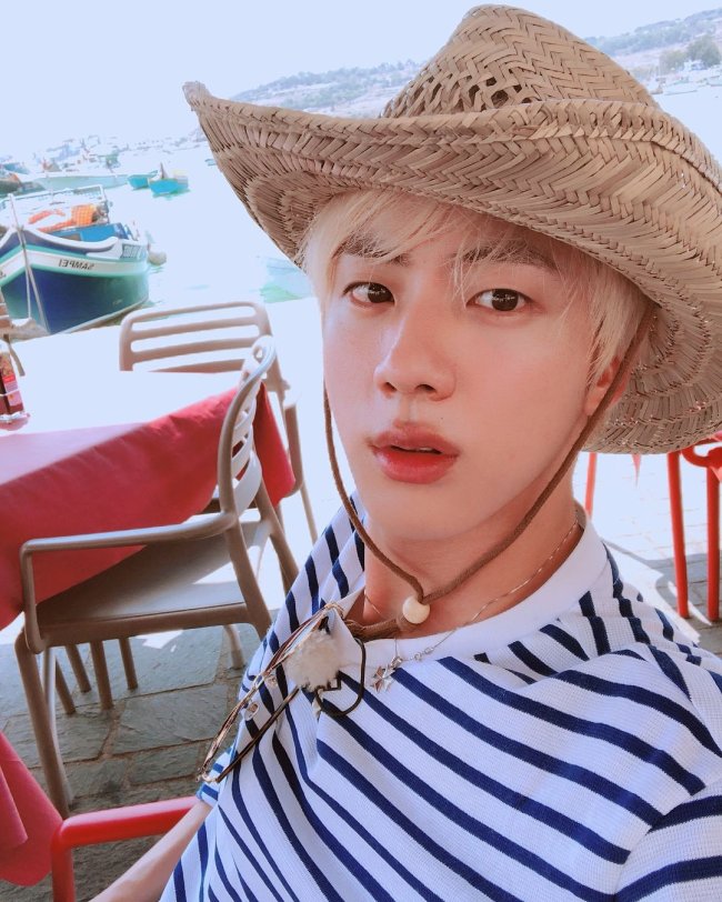 Jin BTS Jin, do grupo de K-pop, BTS sentado em uma cadeira branca de plástico, com expressão séria, usando chapéu de palha, camisa listrada e colares.