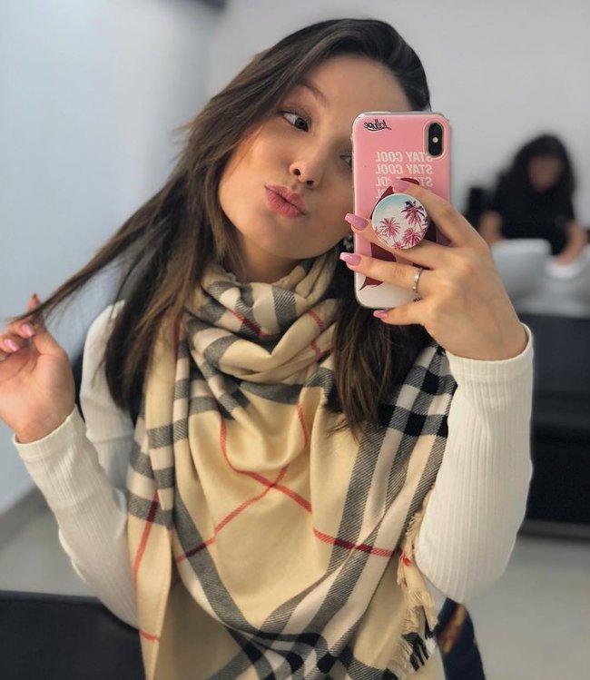 Larissa Manoela com cabelo médio posando para foto fazendo biquinho segurando celular rosa com cachecol xadrez bege e listras preta e vermelho.