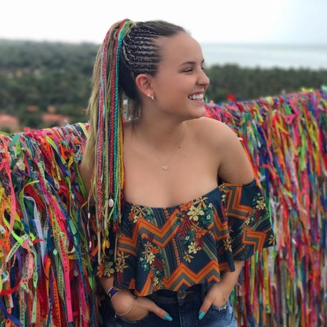 Larissa Manoela trancinha colorida Larissa Manoela com cabelo longo olhando para o lado sorridente posando em frente a muro com fitinhas, usando cropped ciganinha com as mãos no bolso.